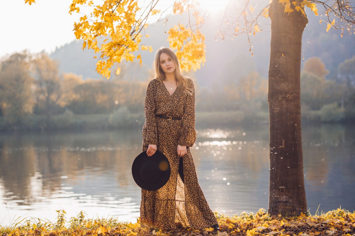 Frau steht mit einem Leo Kleid im boho style am see Ufer neben einem Baum.